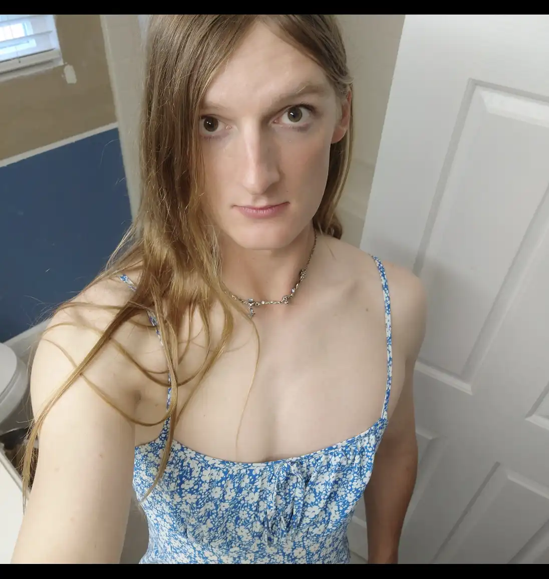 A person with long, wet-looking blonde hair stares intensely at the camera while wearing a blue floral spaghetti strap top…