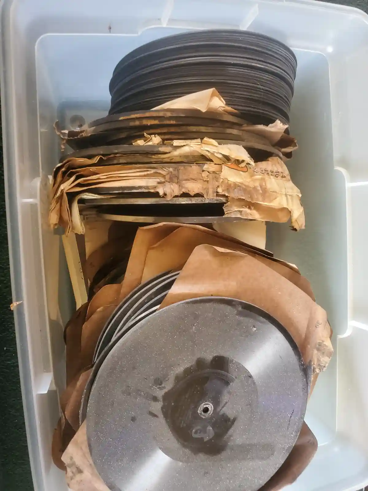 A clear plastic storage bin contains stacks of old, dusty 78 rpm vinyl records, many wrapped in brown paper sleeves that are…