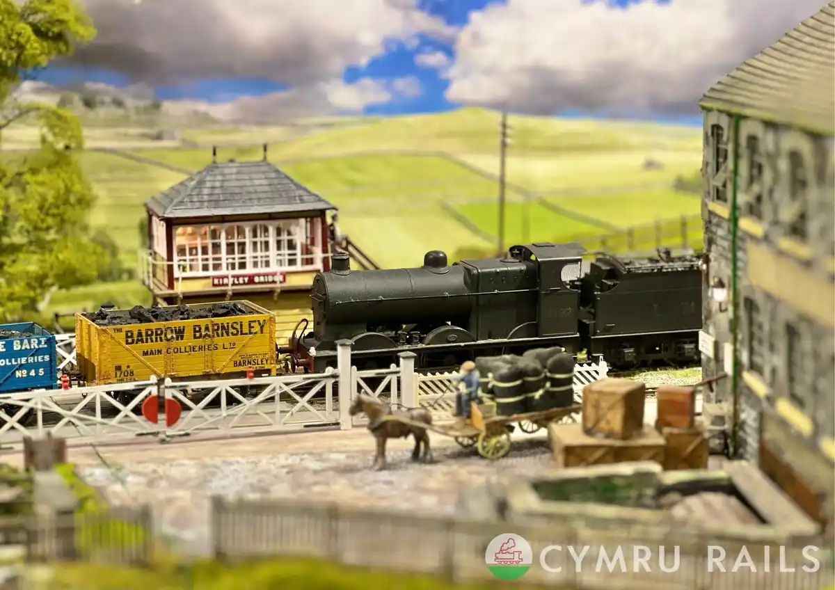 A detailed model railway scene featuring a black steam locomotive pulling coal wagons past a signal box labeled 'KIRKLEY…
