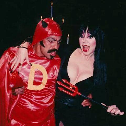 A man in a red devil costume with a 'D' on his chest and horns looks down at a woman in a black devil costume holding a red…