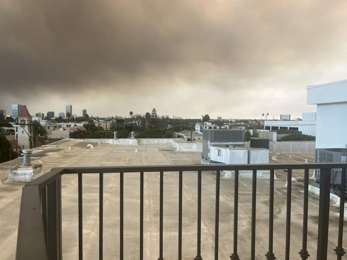 A photo taken from a rooftop balcony shows a cityscape under a thick, ominous brown haze that obscures the sky and distant…