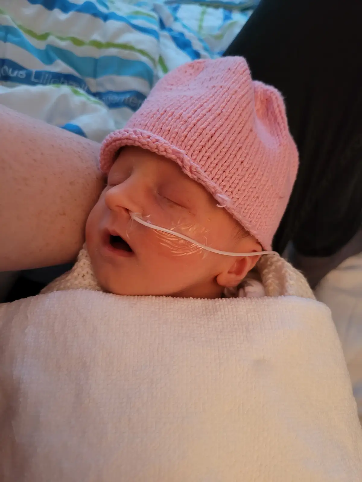 A newborn baby, wrapped in a white blanket and wearing a pink knit hat, sleeps peacefully while an oxygen tube is inserted…