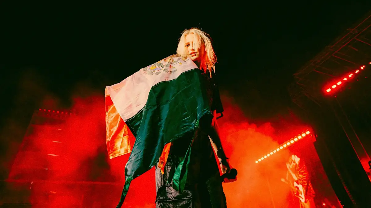 A blonde woman stands on a concert stage draped in a Mexican flag, holding a microphone, with red stage lights and smoke in…