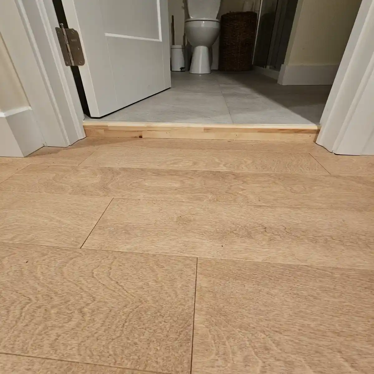 A low-angle photo shows a light wood-patterned floor leading to a bathroom doorway. The bathroom floor is visibly…