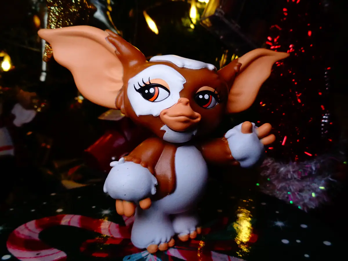 A small brown and white Gremlin toy figure with large ears and expressive eyes stands in front of a decorated Christmas tree…