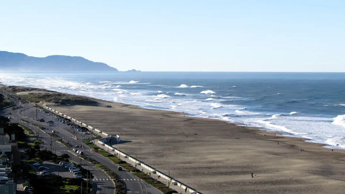 A wide-angle photo of a coastal highway running parallel to a sandy beach with ocean waves breaking in the distance. The…