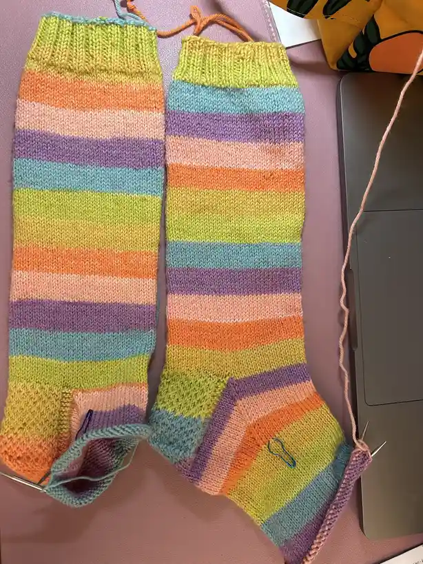Two mismatched, colorful striped socks are shown mid-knitting on a pink surface next to a laptop, with the text 'they're not…