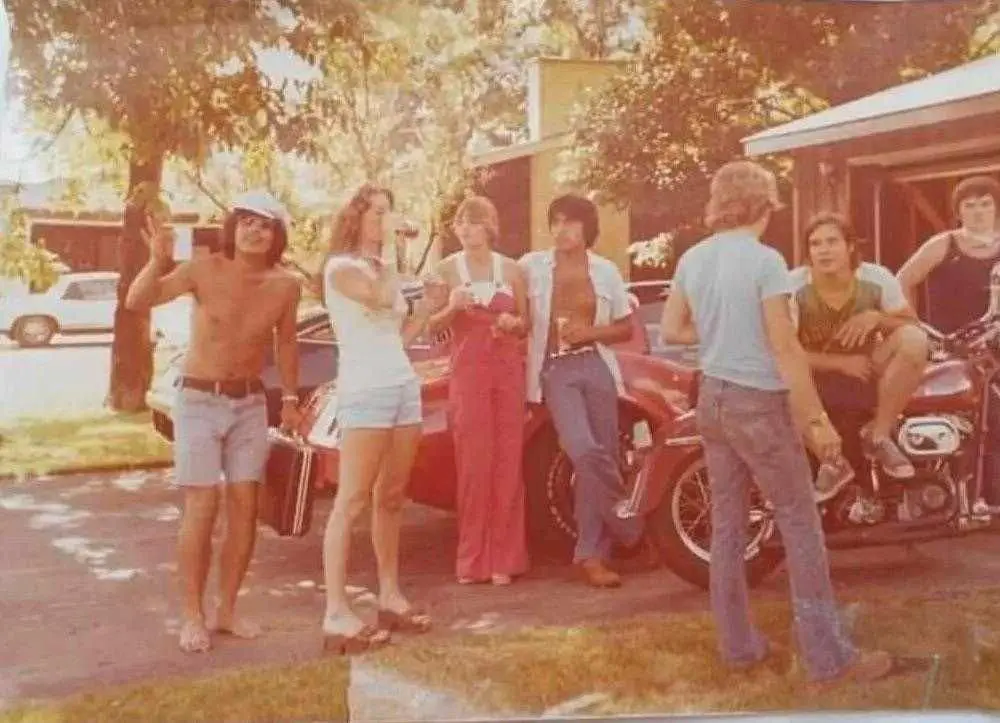 A faded, sun-bleached 1970s photo shows a group of young adults in casual, era-specific clothing gathered around a red car…