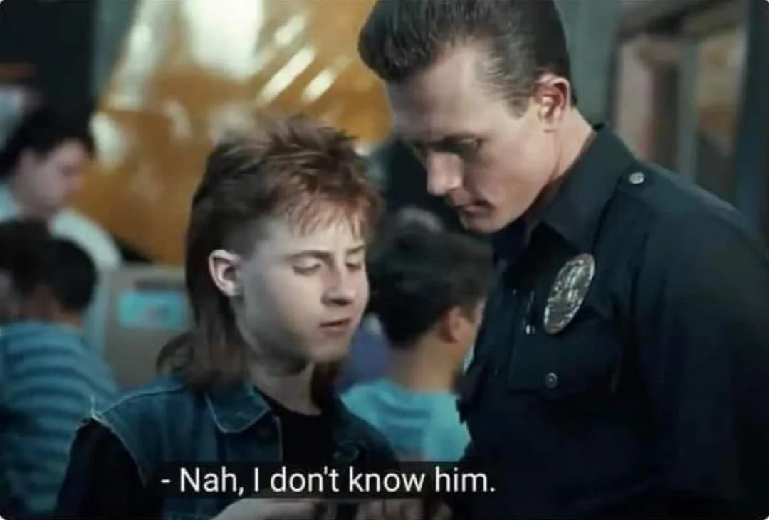 A still from a movie shows a young man with mullet hair wearing a denim vest looking down while a police officer in uniform…