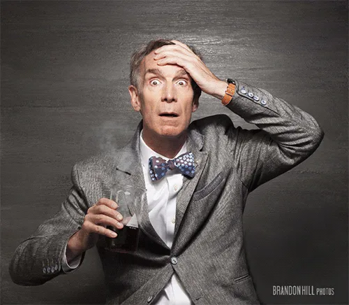 A man in a grey suit and patterned bow tie holds a steaming glass of dark liquid while clutching his head with one hand, his…
