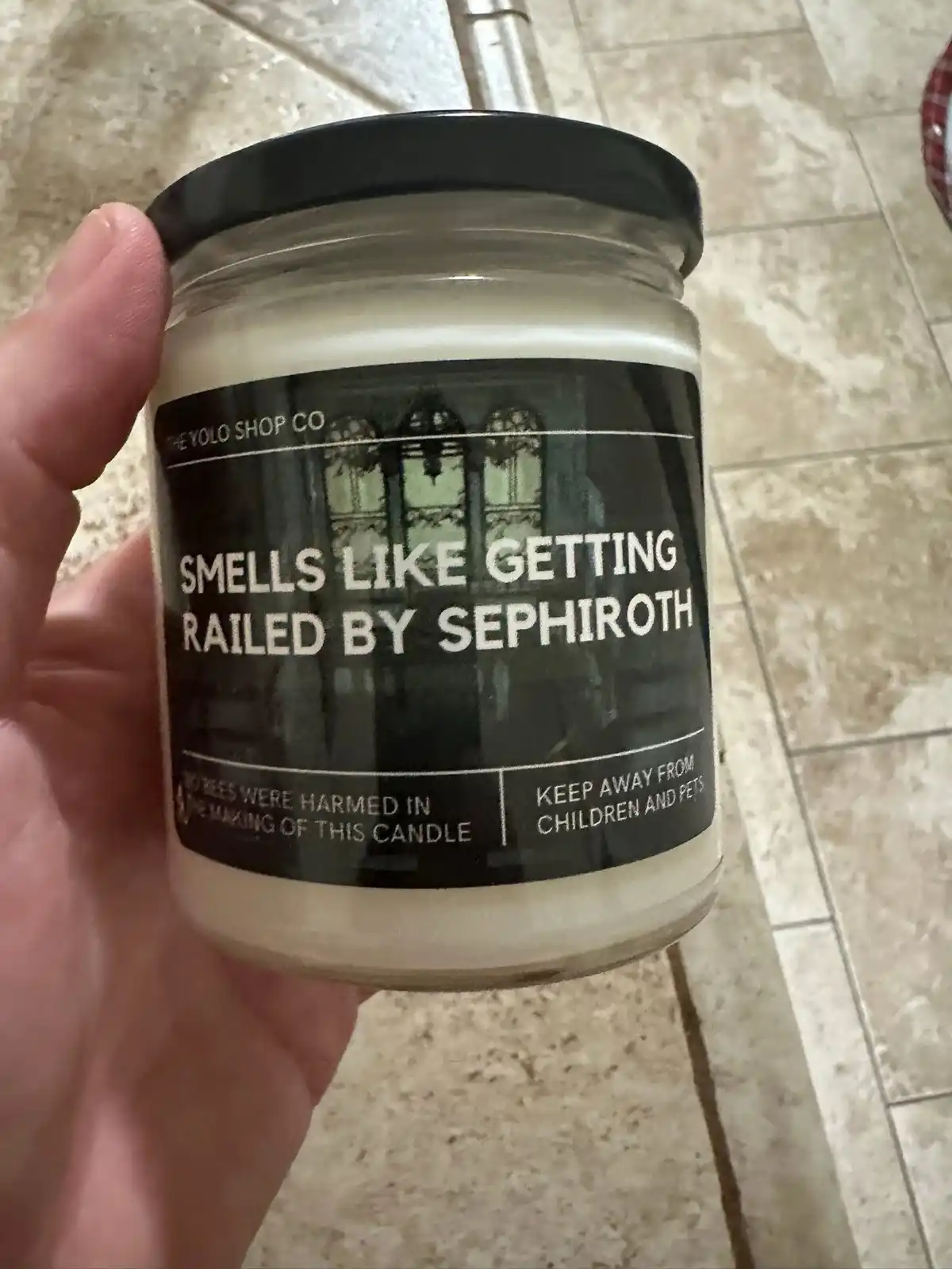 A hand holds a glass jar candle with a black label that reads 'SMELLS LIKE GETTING RAILED BY SEPHIROTH' and includes smaller…
