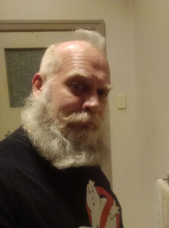 A man with a full white beard and mustache stares directly at the camera with a serious expression, wearing a black t-shirt…