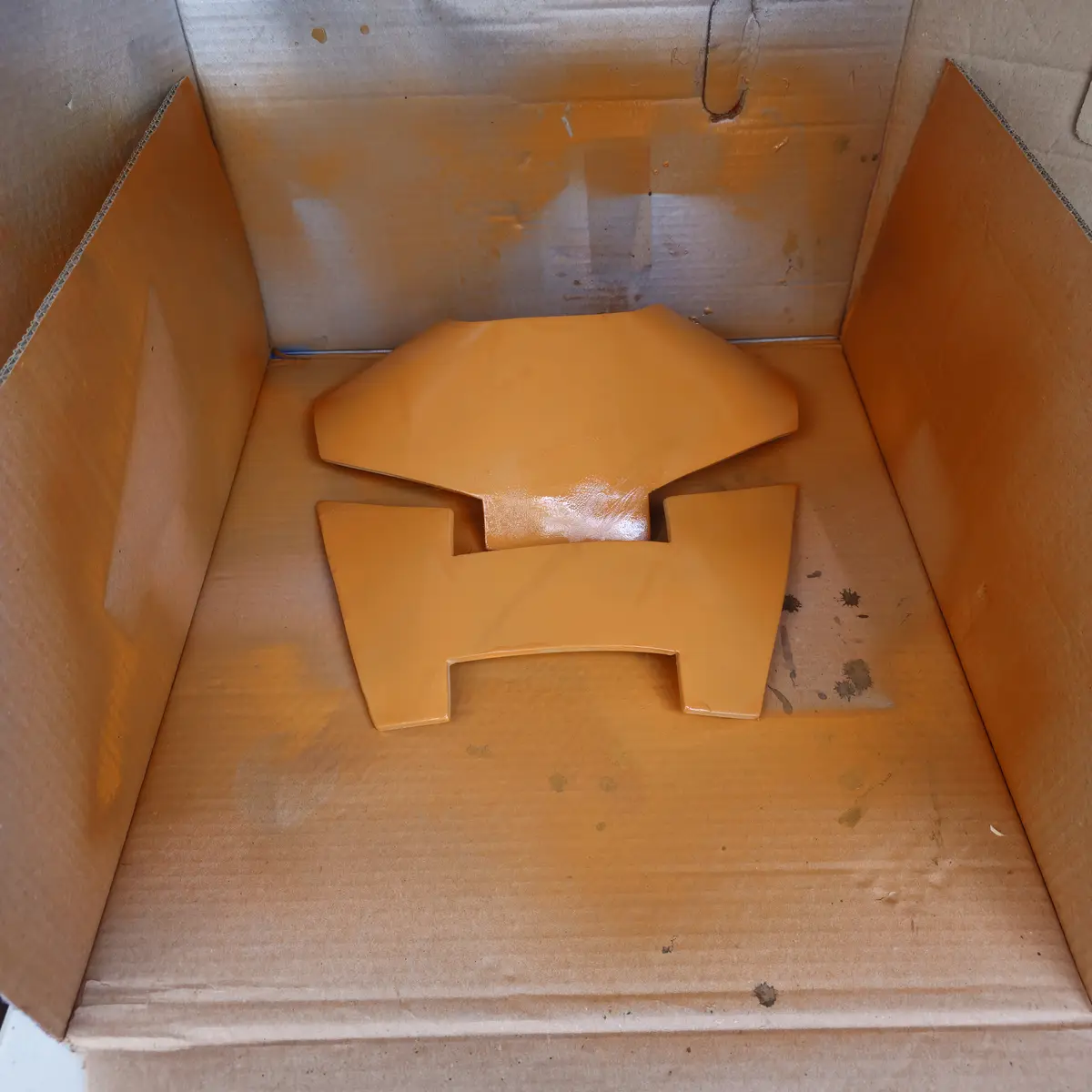 A top-down view of two freshly spray-painted orange car parts resting inside a cardboard box, with orange paint splatters…
