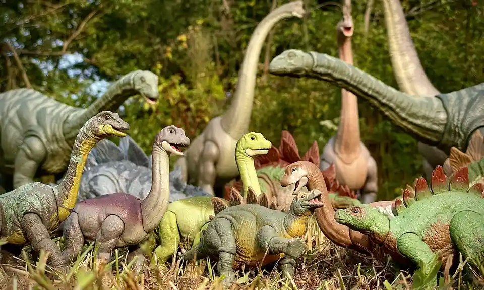 A group of plastic dinosaur toys, including long-necked sauropods and spiky stegosaurs, are arranged outdoors in grass and…