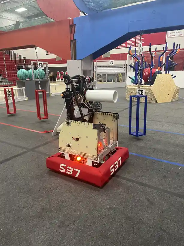 A red robot with the number 537 on its base is shown in a gymnasium during what appears to be a robotics competition. The…