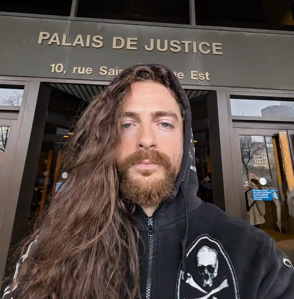 A man with long brown hair and a beard, wearing a black hoodie with a skull and crossbones design, takes a selfie in front…