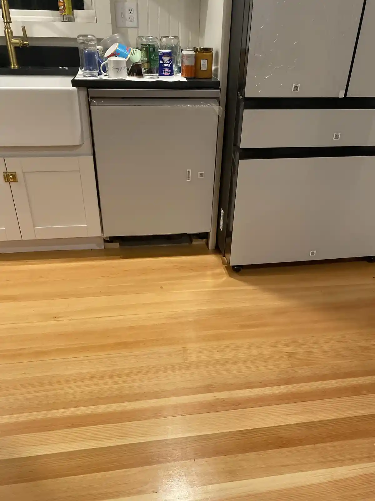 A low-angle shot of a clean kitchen with light wood flooring, a white sink, and a stainless steel refrigerator. On the…