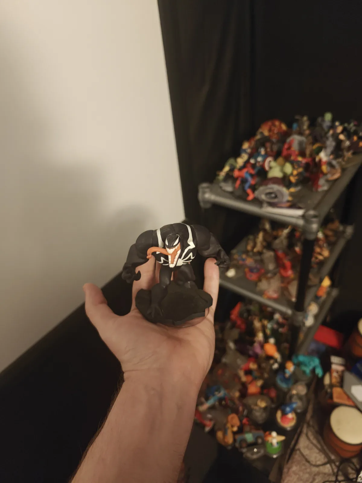 A person holds a Venom action figure in their hand, with a metal shelving unit filled with numerous other action figures…