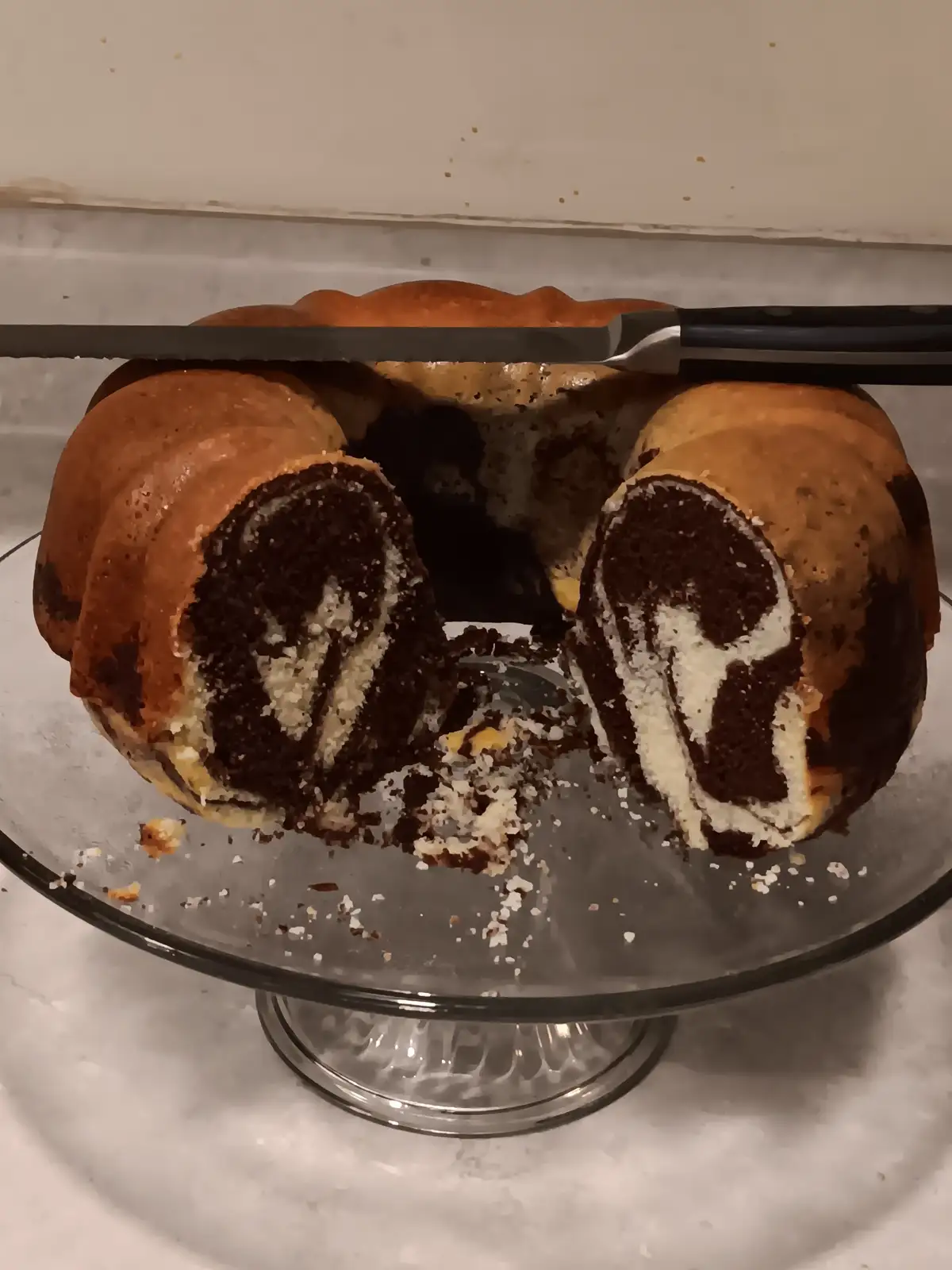 A close-up photo shows a partially sliced chocolate and vanilla marble bundt cake on a glass cake stand, with a serrated…