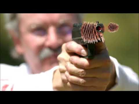 A man with a mustache holds a handgun pointed forward, but the barrel is replaced with a pine cone, and another pine cone is…