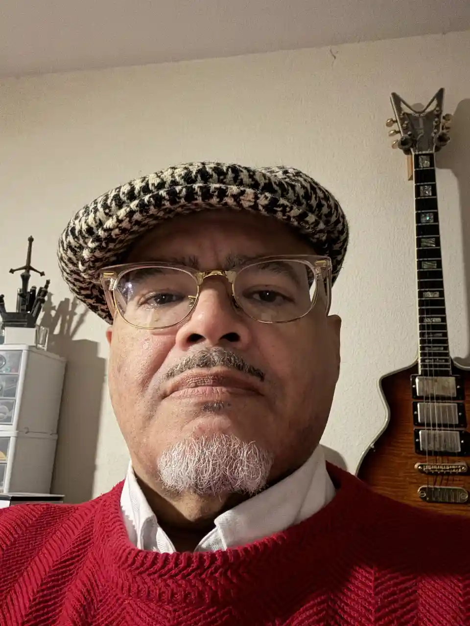 A close-up selfie of a middle-aged man with a serious, unimpressed expression, wearing a black-and-white houndstooth flat…