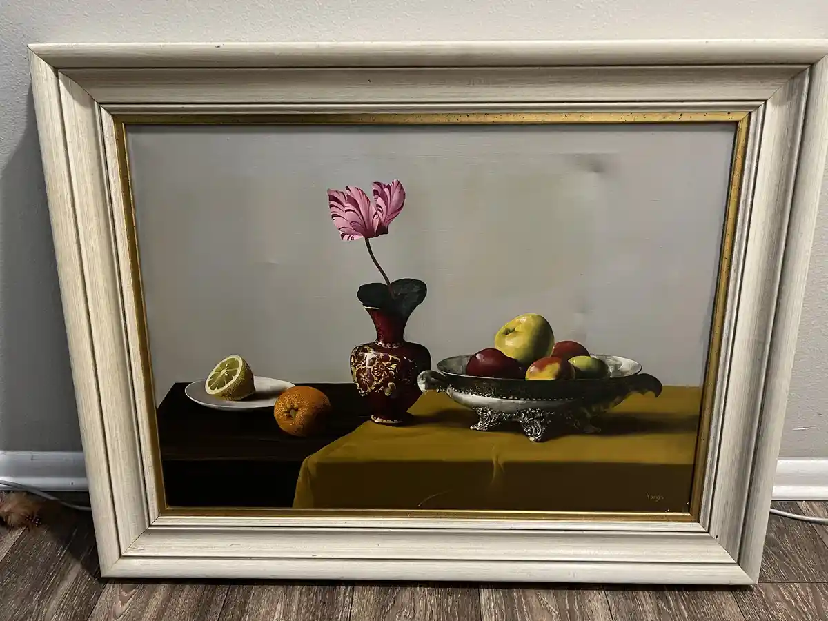 A framed still life painting depicts a red vase with a pink flower, a silver bowl of fruit, a sliced lemon on a plate, and…
