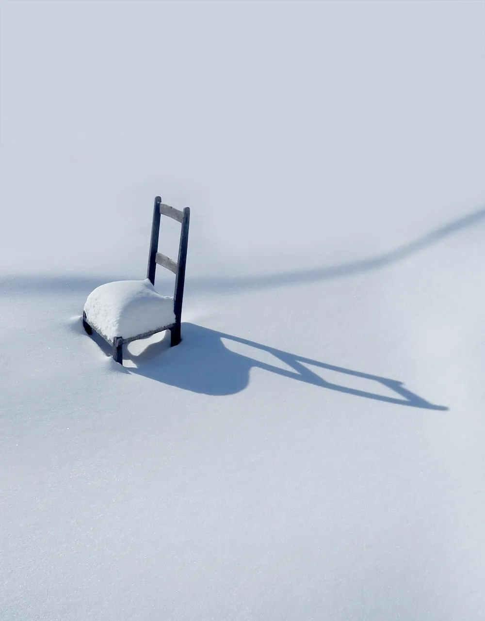 A solitary wooden chair sits partially buried in deep snow under bright sunlight, casting a long, sharp shadow across the…