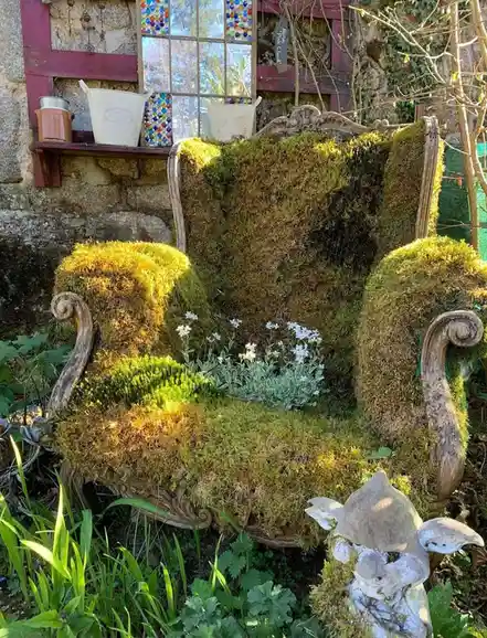 An ornate, antique-style wooden chair is completely covered in thick, vibrant green moss, with small white flowers growing…