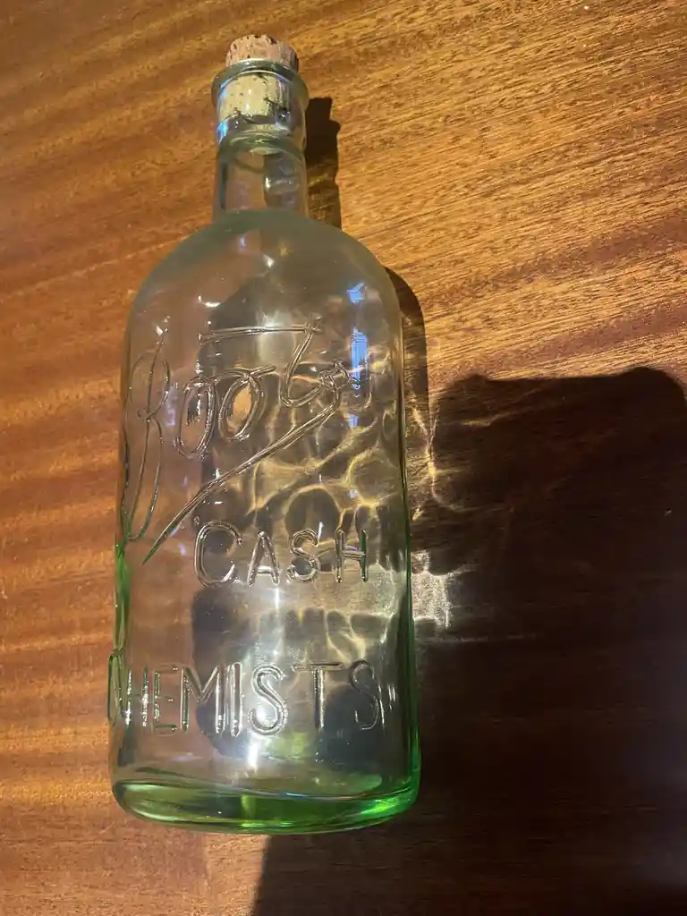 A vintage green glass bottle with embossed text reading 'Roots Cash Chemists' sits on a wooden surface, sealed with a cork…