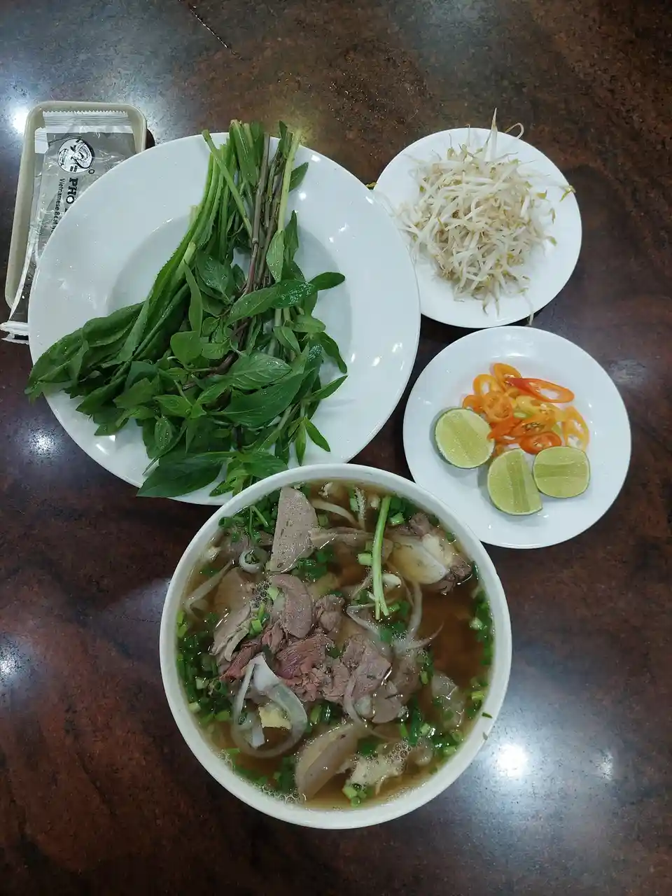 A top-down photo of a bowl of Vietnamese pho with meat, noodles, and green onions, surrounded by plates of fresh herbs, bean…
