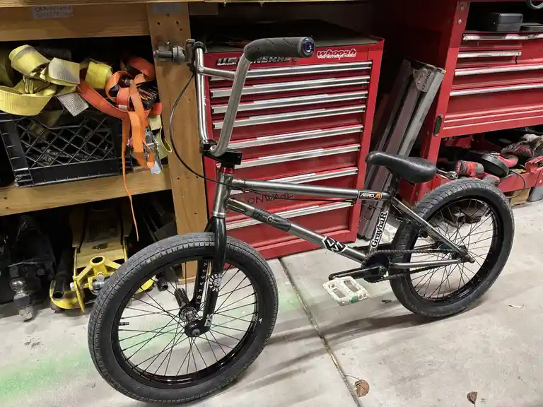 A silver and black BMX bike is parked in a cluttered garage next to red toolboxes, with yellow and orange tie-down straps…