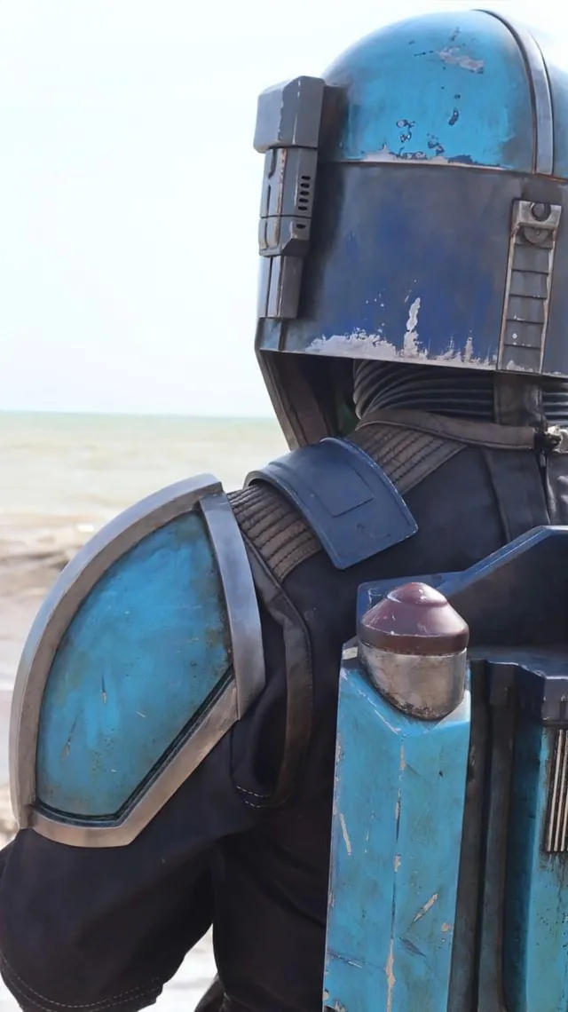 A person in a blue and black Mandalorian armor costume stands with their back to the camera, facing a beach and ocean. The…