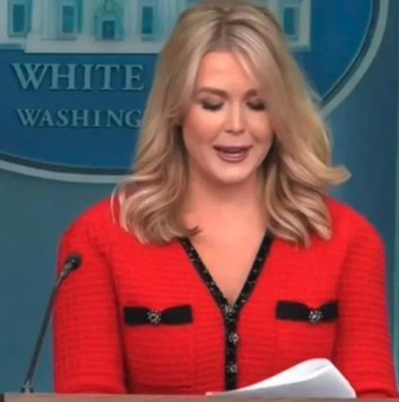 A woman with blonde hair wearing a red jacket stands at a podium in front of a blue backdrop that reads 'WHITE WASHING'. She…