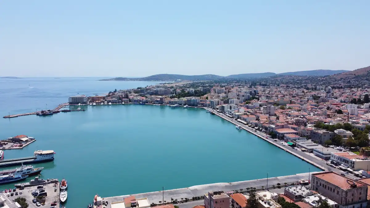 An aerial view of a coastal town with a large harbor filled with turquoise water, surrounded by buildings with red-tiled…