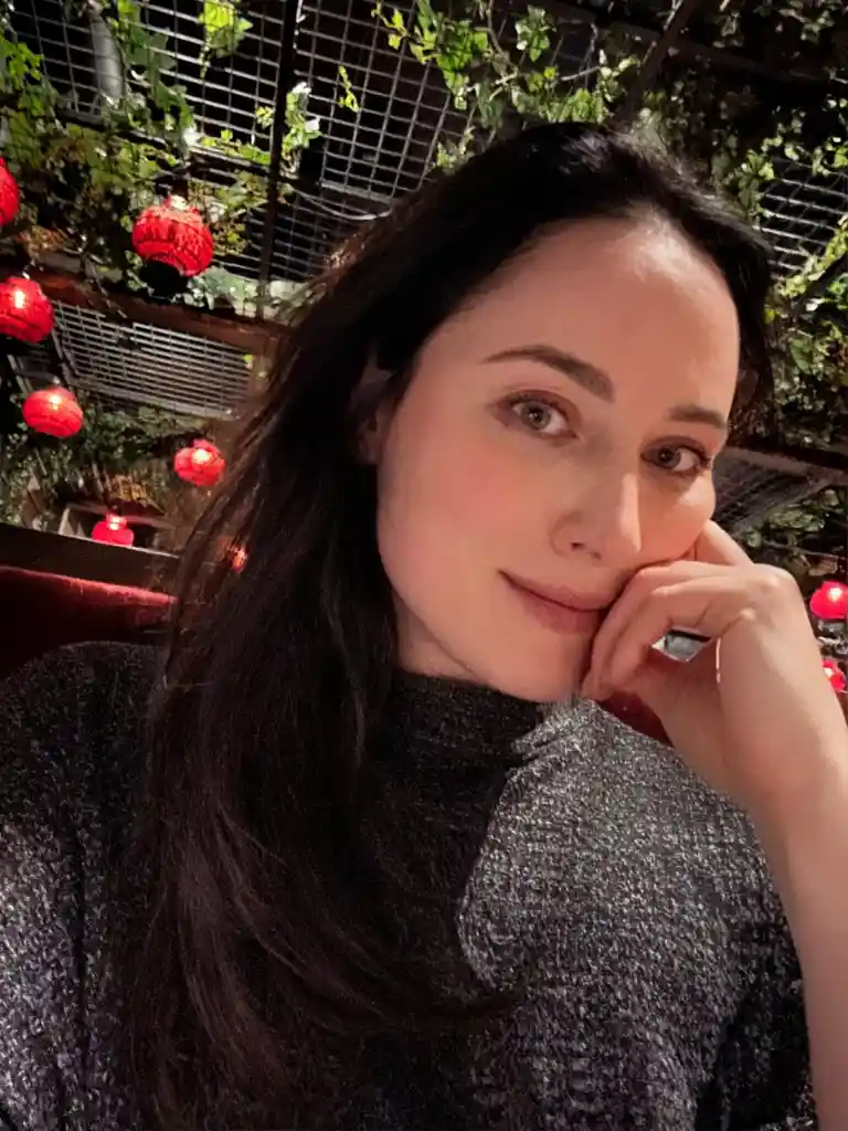 A woman with long dark hair and a subtle smile rests her chin on her hand, looking directly at the camera. She wears a dark…