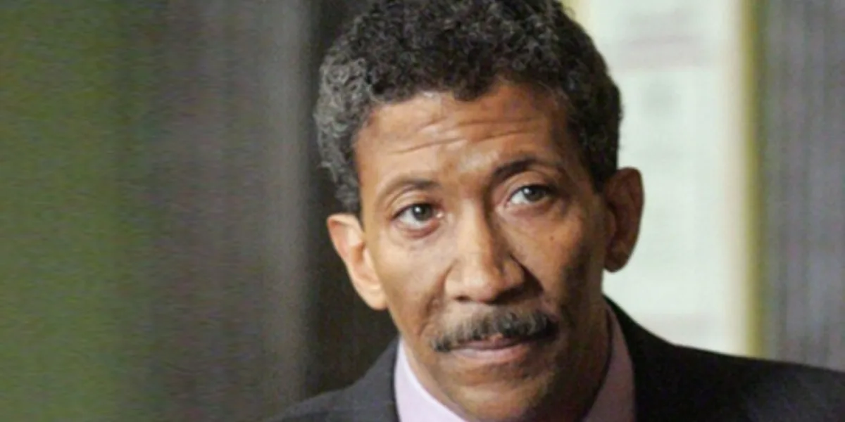A close-up photo of a man with short curly hair and a mustache, wearing a suit and tie, looking slightly upward with a…
