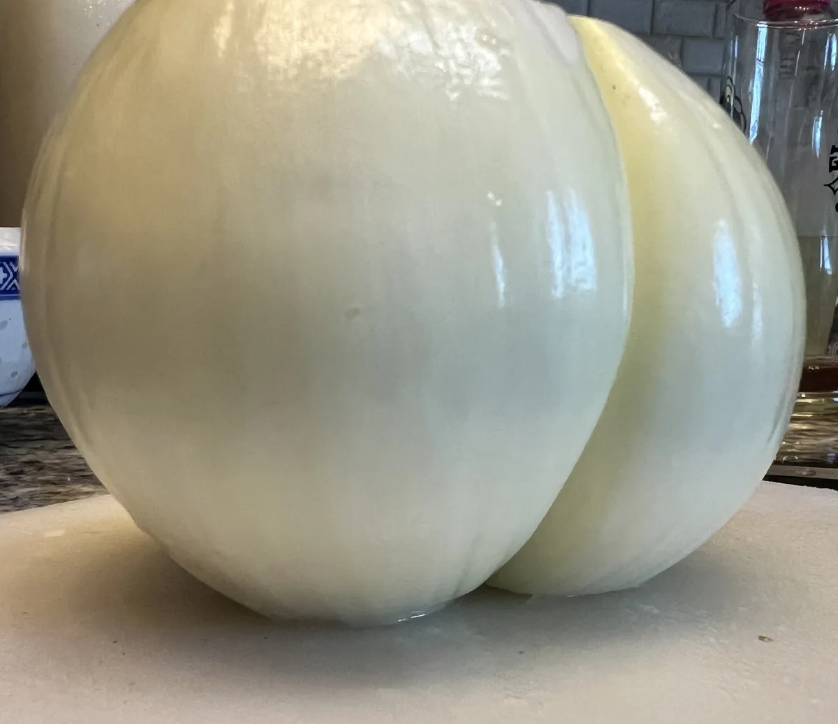 A close-up photo shows a very large, peeled white onion sitting on a cutting board, its bulbous shape and smooth, pale skin…