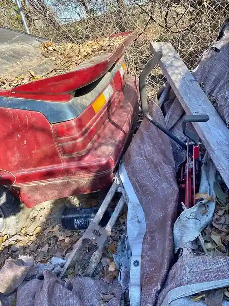 A red car's rear end is partially buried in a pile of debris including wooden planks, tarpaulins, and leaves, next to a…