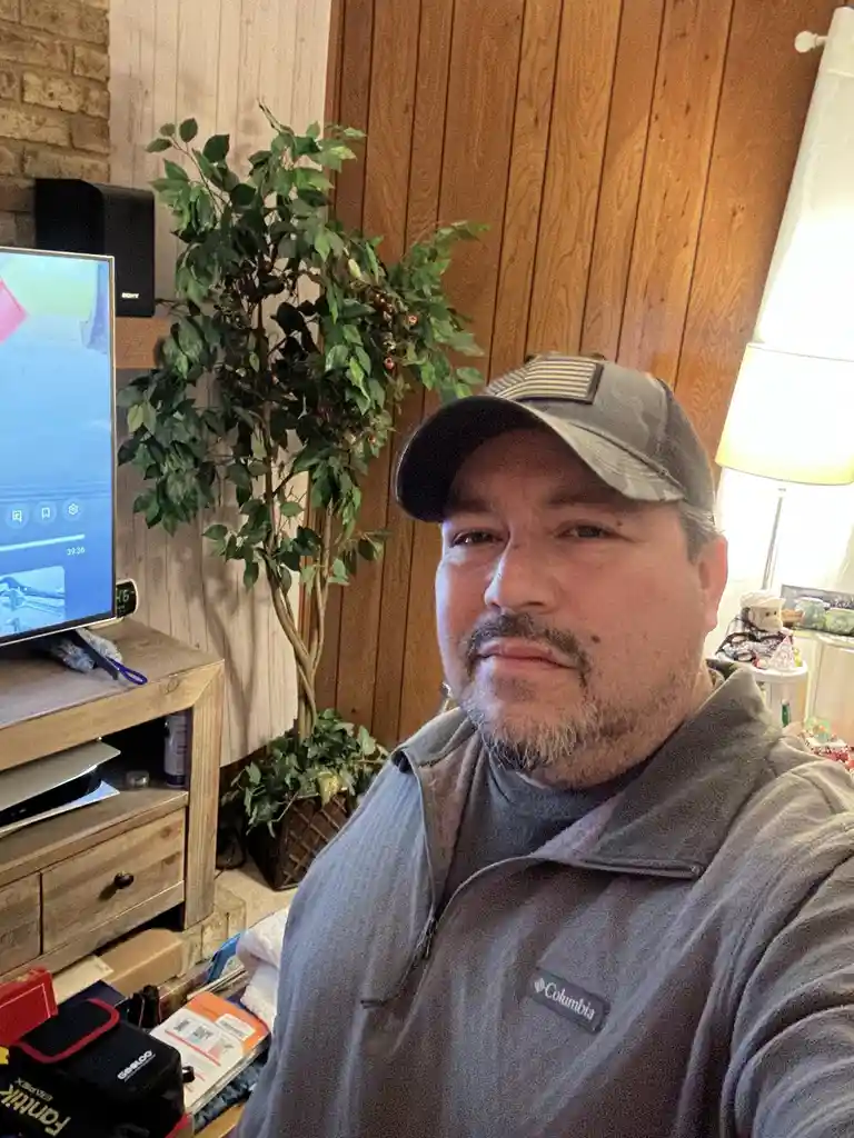 A man in a grey Columbia vest and an American flag baseball cap stares directly at the camera with a neutral expression…