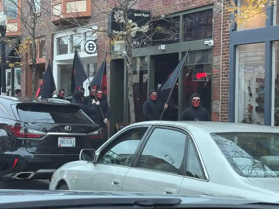 A group of people wearing black balaclavas and red skull-patterned face coverings stand outside a restaurant holding black…
