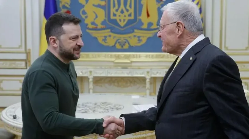 Two men are shaking hands in a formal room with a Ukrainian coat of arms behind them. The man on the left, wearing a dark…