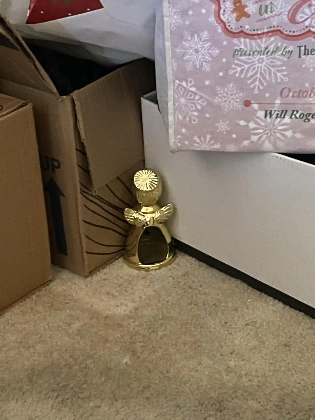 A small, golden angel figurine with a hollow body sits on a carpeted floor in a corner, surrounded by cardboard boxes and a…
