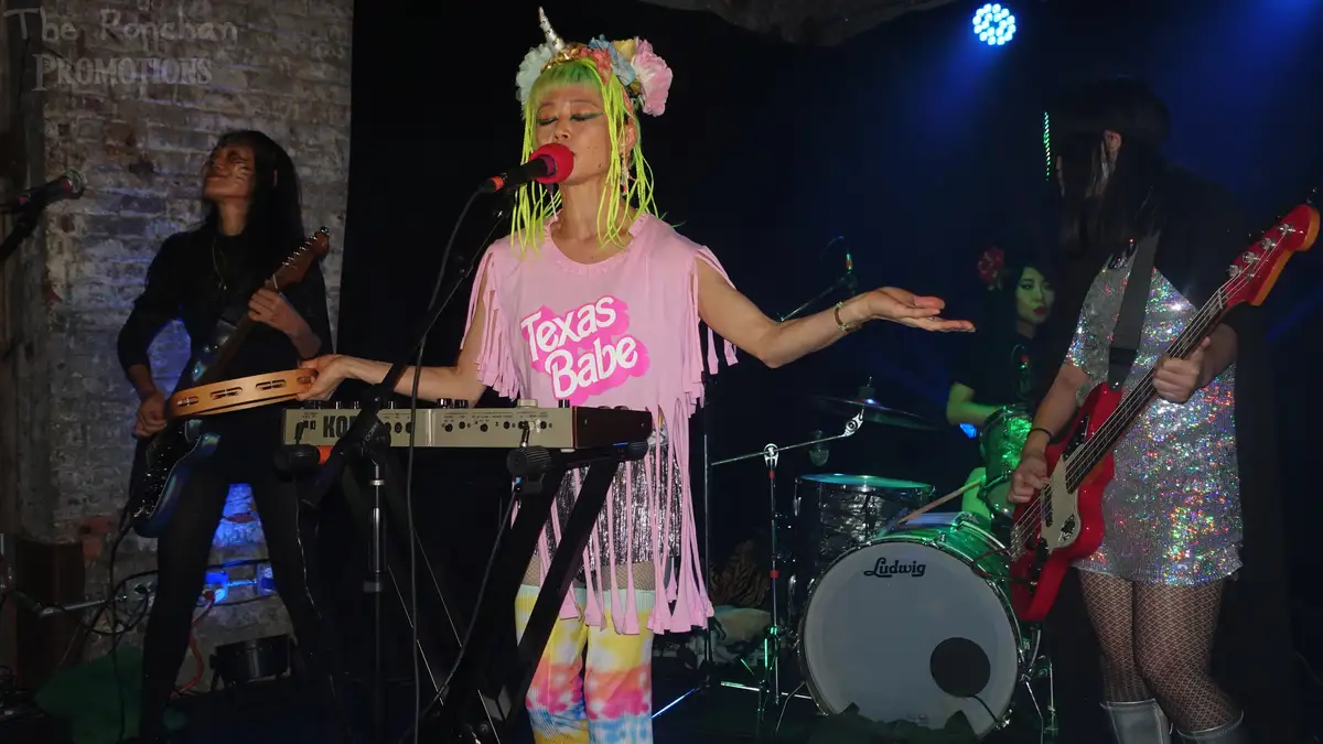A band performs on stage with a singer in a pink 'Texas Babe' shirt and neon green hair wearing a floral crown, singing into…