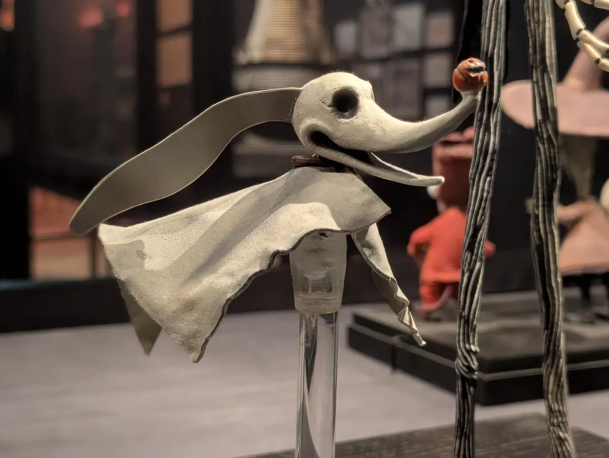 A stop-motion animation puppet of a skeletal dog with a long snout, wearing a white cape, is displayed on a glass stand. It…