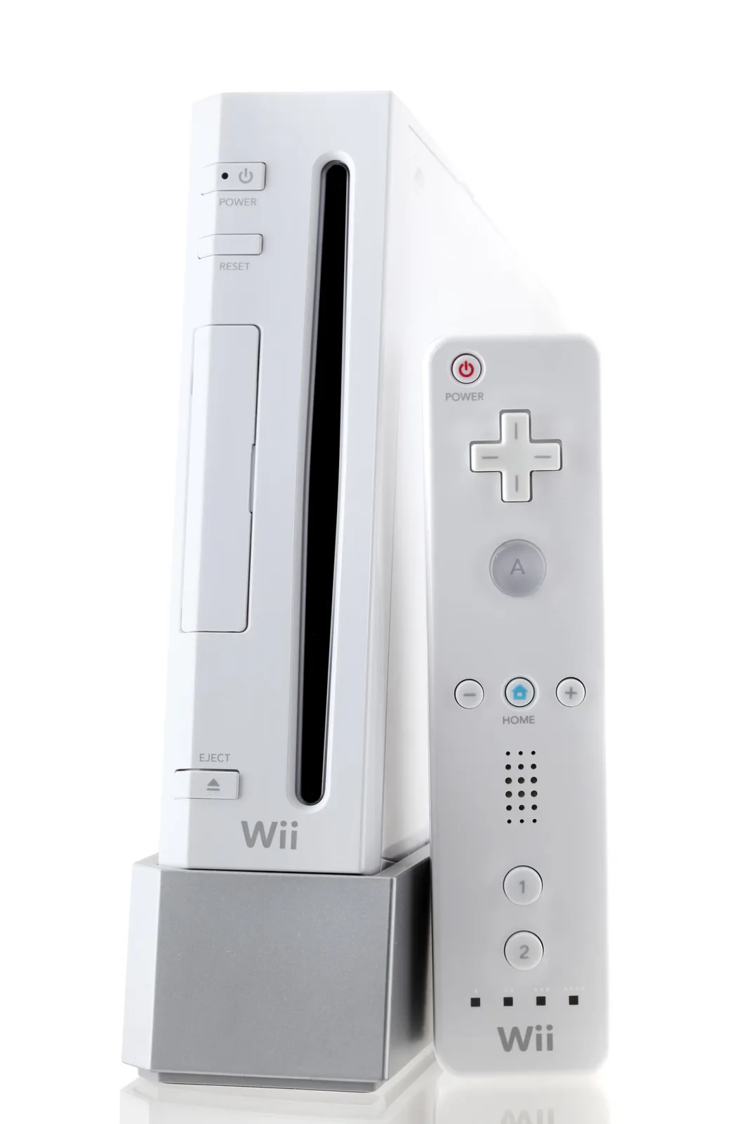 A white Nintendo Wii console sits upright in its silver stand next to a white Wii Remote controller, both against a plain…