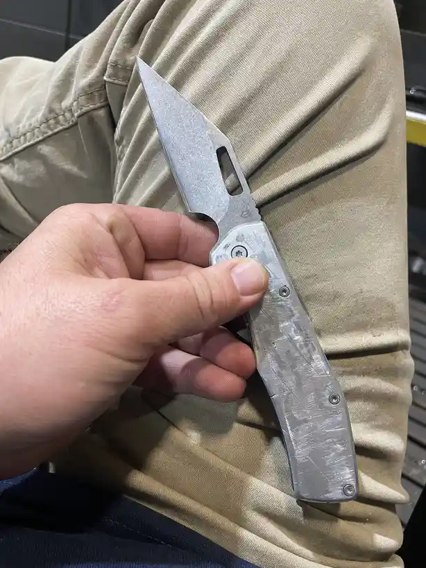 A person's hand holds a silver, worn-looking folding knife with a sharp blade and visible screws, resting against their…