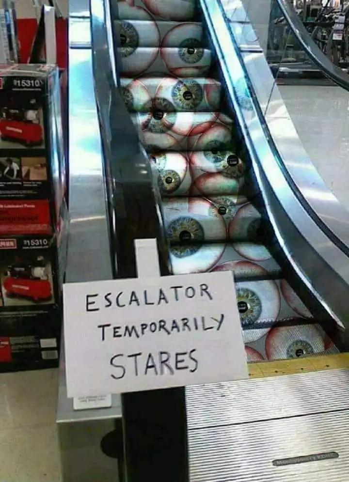 An escalator with steps covered in printed images of human eyeballs is shown, with a handwritten sign in front that reads…