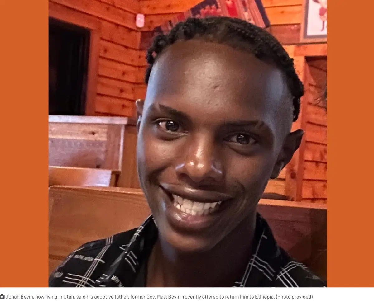 A close-up selfie of a smiling young Black man with short braided hair, wearing a black and white plaid shirt, set against a…