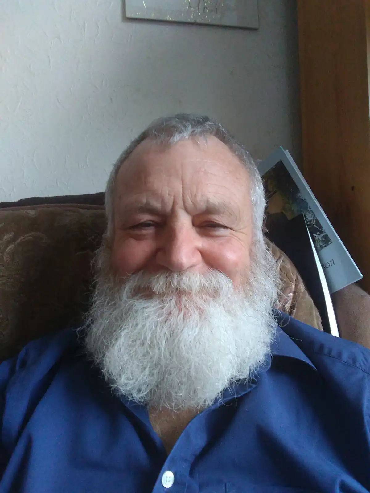 An older man with a long white beard and gray hair is smiling while seated in a brown armchair, wearing a blue button-up…