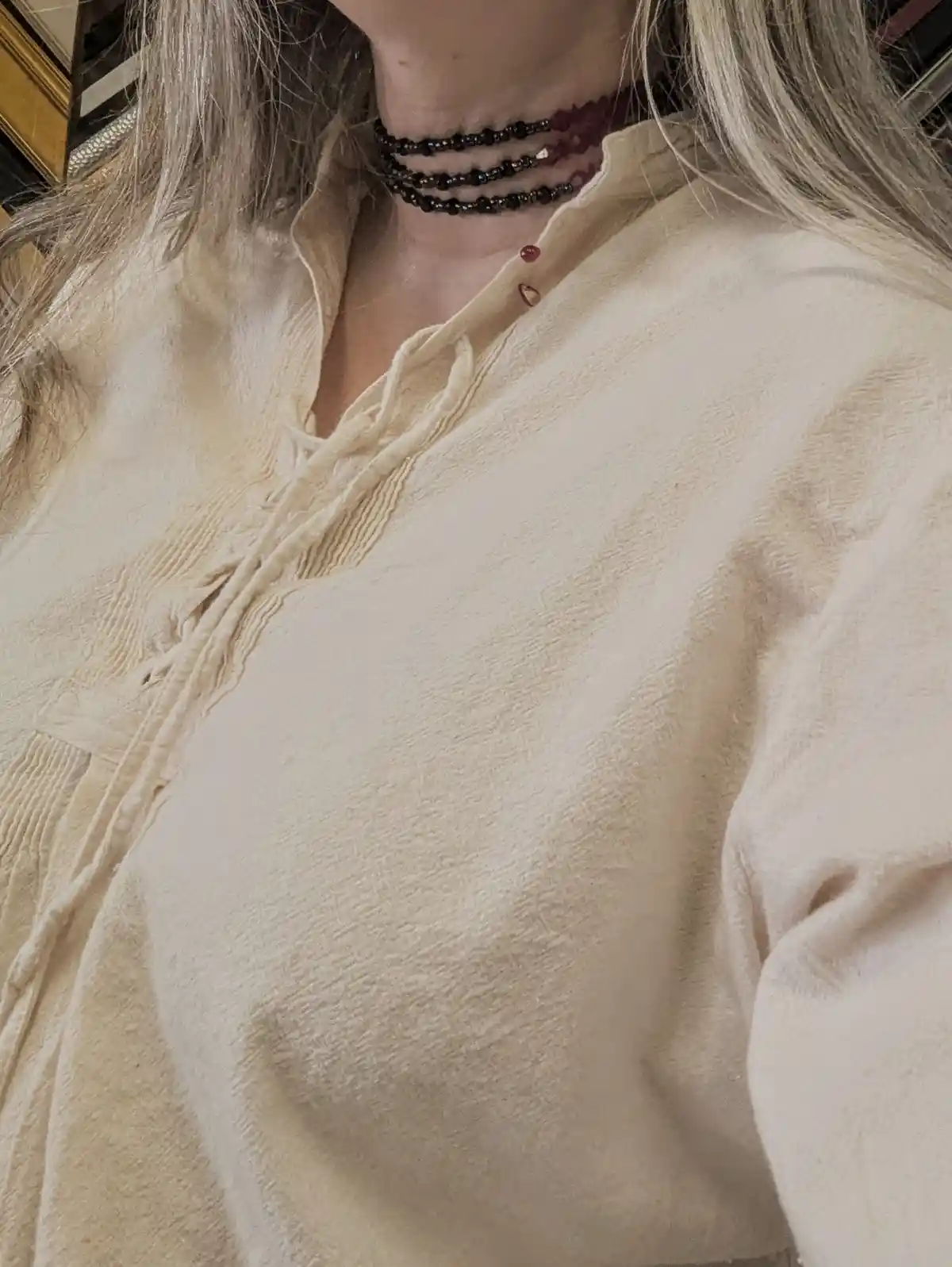 A close-up selfie shows a person with blonde hair wearing a cream-colored textured shirt with a laced front and a…